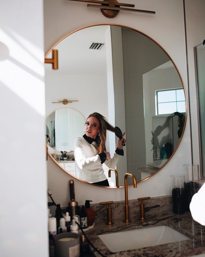 Contact A young woman brushes her long hair, reflected in a round mirror in an elegant bathroom setting.