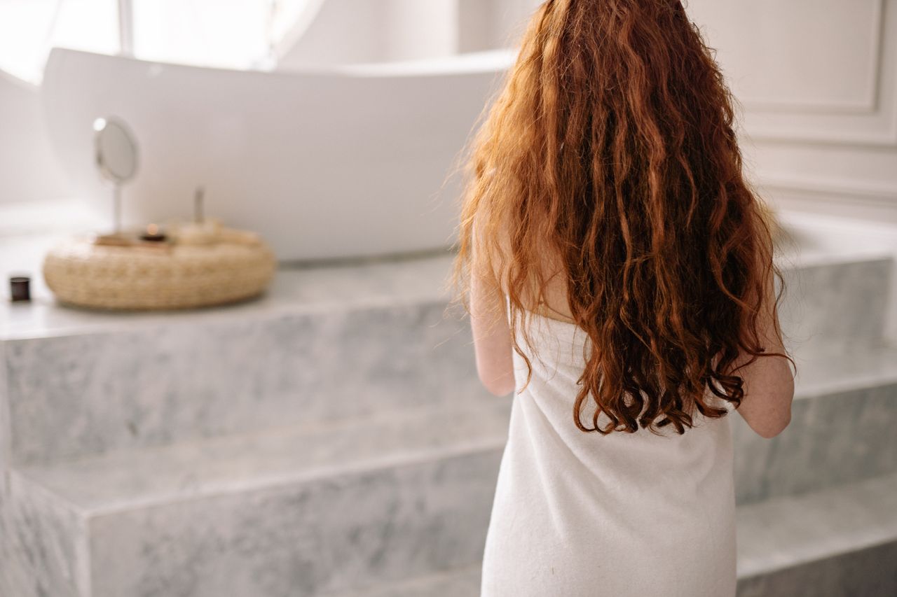 A woman with long red hair wrapped in a towel stands near a bathtub, embodying relaxation and self-care.