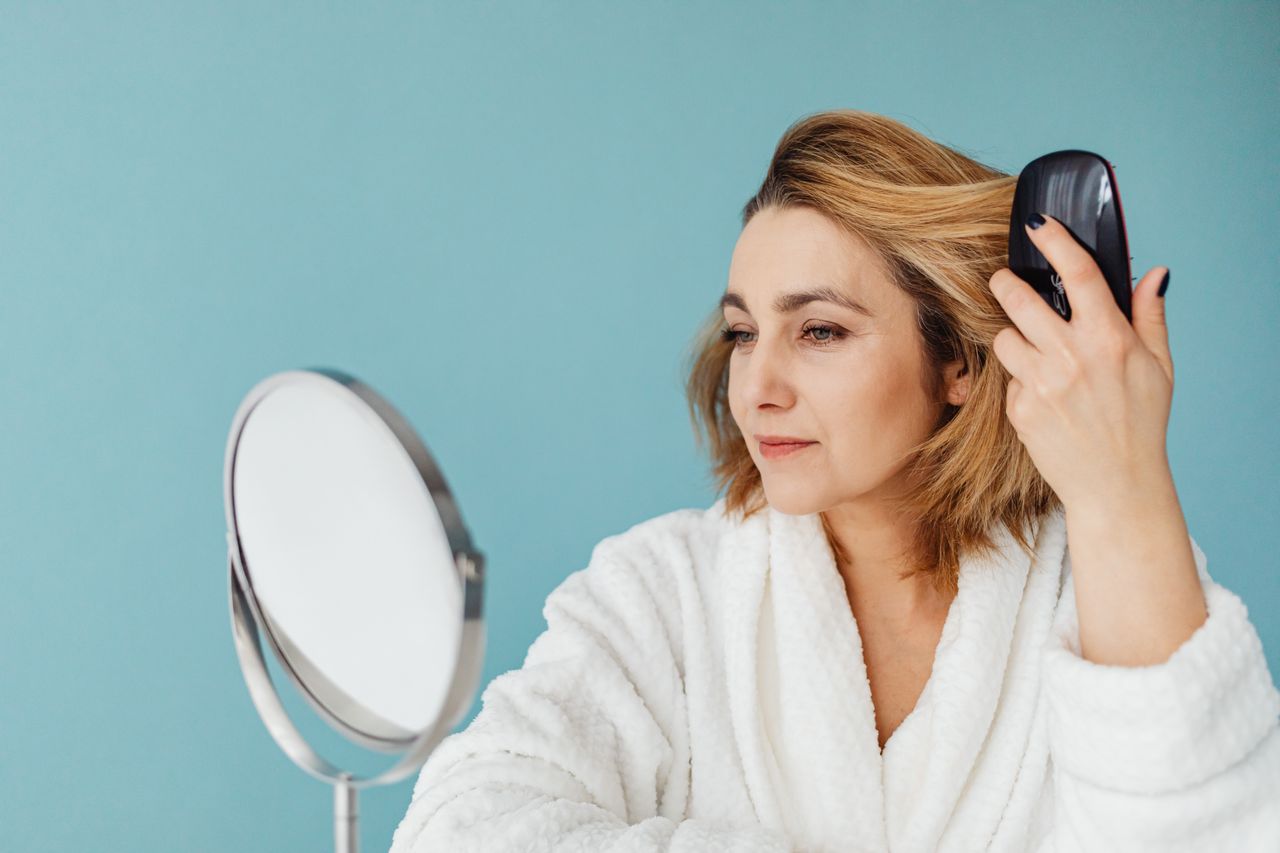 A woman in a bathrobe combs her hair while looking into a mirror with a calm expression.